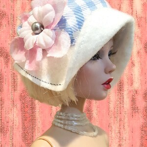 K&ouml;nnte beinhalten: Eine Puppe tr&auml;gt einen blau-wei&szlig; karierten Hut mit breiter wei&szlig;er Krempe, verziert mit einer gro&szlig;en hellrosa Stoffblume und einer grauen Perle in der Mitte. Die Puppe hat blondes Haar, eine Perlenkette und ein blaues Oberteil.