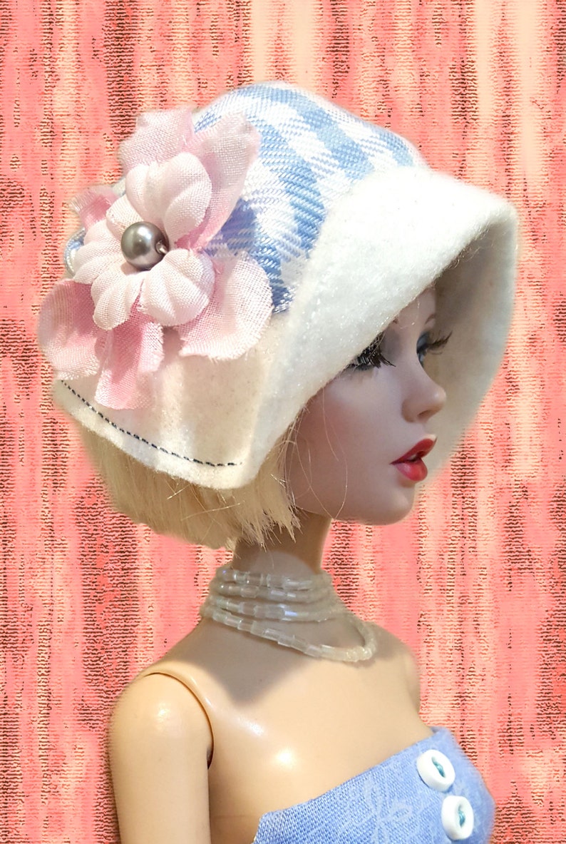 K&ouml;nnte beinhalten: Eine Puppe tr&auml;gt einen blau-wei&szlig; karierten Hut mit breiter wei&szlig;er Krempe, verziert mit einer gro&szlig;en hellrosa Stoffblume und einer grauen Perle in der Mitte. Die Puppe hat blondes Haar, eine Perlenkette und ein blaues Oberteil.