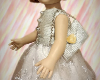 Könnte beinhalten: Puppe mit rotbraunem Haar und blauen Augen, gekleidet in einem silbernen Kleid mit transparentem Überzug und passender Schleife. Die Puppe trägt eine kleine Handtasche und schwarze Schuhe. Das Kleid ist mit silbernen Verzierungen versehen.