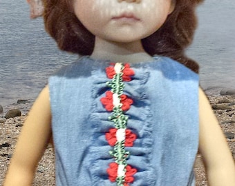 May include: A doll wearing a denim blue dress with red floral accents and a matching floral hat. The dress features a ruffled bodice with red flower embellishments. The hat has a straw brim and is decorated with red flowers and green leaves.