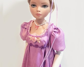 May include: A doll wearing a purple dress with puff sleeves, a pearl necklace, and a floral headband. The doll has a delicate face with blue eyes and is posed against a white background. Ribbons cascade down from the headband.