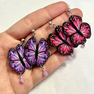 Puede incluir: Un par de pendientes colgantes con diseños de mariposas. Las mariposas están hechas de arcilla polimérica y tienen detalles intrincados. Un par es rosa y el otro es morado.