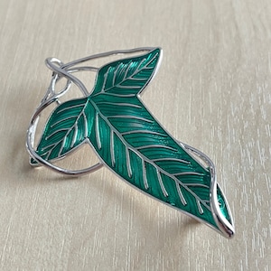 May include: A silver-toned metal leaf-shaped brooch with a vibrant green enamel finish. The brooch features detailed veining and a delicate, swirling frame, creating an elegant accessory. The leaf design is a classic nature-inspired motif.