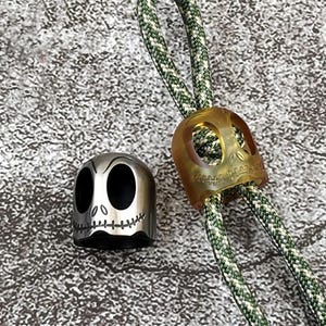 May include: Two skull-shaped zipper pulls, one is silver and the other is translucent yellow. Both are attached to a green and white braided cord.