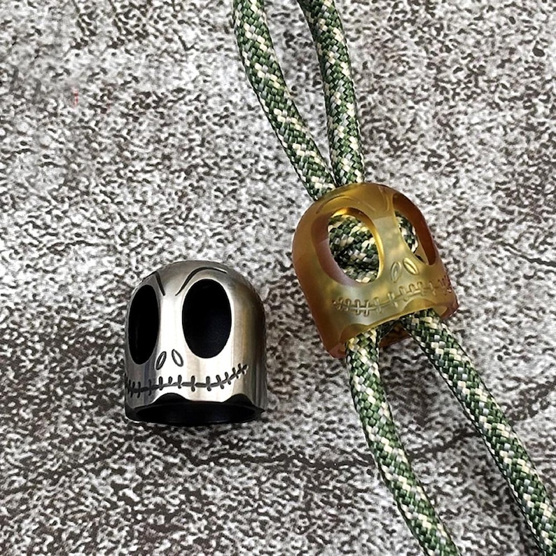 May include: Two skull-shaped zipper pulls, one is silver and the other is translucent yellow. Both are attached to a green and white braided cord.