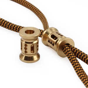 May include: Two gold-colored metal beads with intricate designs and multiple holes. The beads are threaded onto a black and yellow braided cord.
