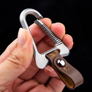 Op de afbeelding: Een zilverkleurige karabijnhaak met een bruin lederen riem. De karabijnhaak heeft een uniek ontwerp met een geribbeld gedeelte om te openen. De leren riem is bevestigd met een zilveren klinknagel.