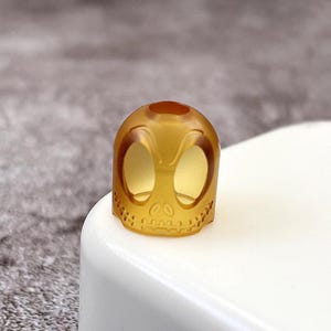 May include: A translucent amber-coloured skull-shaped guitar pick. The pick has two large eye holes and a detailed skull design.