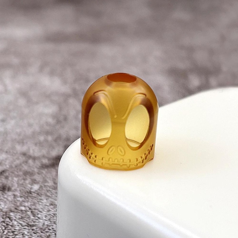 May include: A translucent amber-coloured skull-shaped guitar pick. The pick has two large eye holes and a detailed skull design.