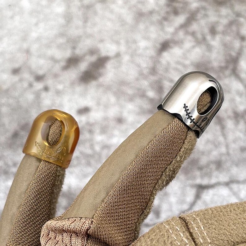 May include: Two fingertip covers, one is tan and the other is silver with a stitched-on design resembling a face. Both are worn on a tan glove.