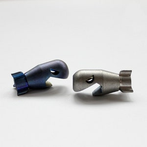May include: Two metal whale-shaped bottle stoppers. One is a dark blue with a gradient effect, and the other is silver. Both have a fin-like tail and a curved opening for the bottle.