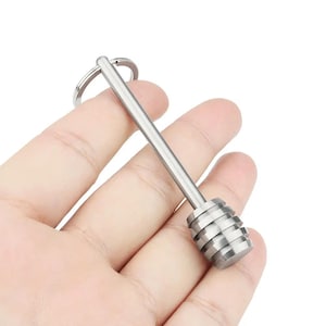 Könnte beinhalten: Ein silberner Honiglöffel aus Metall mit einem Schlüsselanhänger. Der Löffel hat einen langen, dünnen Stiel und einen runden, geriffelten Kopf.