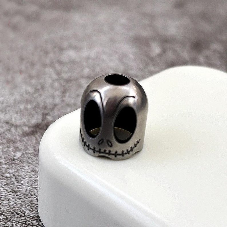 May include: A silver-coloured metal skull charm with a stitched mouth and hollow eye sockets. The charm is sitting on a white surface.