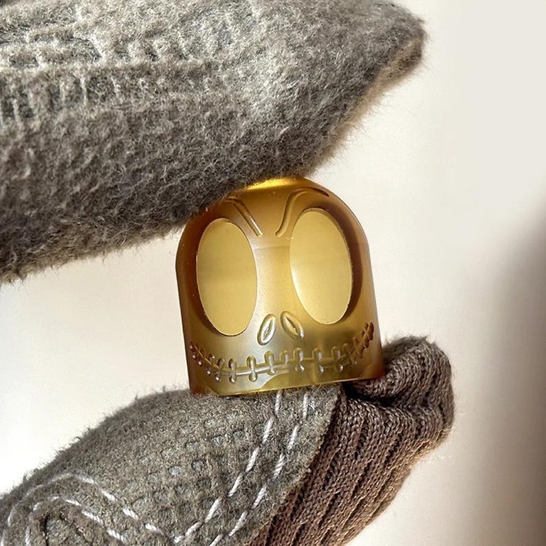 May include: A gold-coloured bottle stopper featuring a detailed skull design. The skull has two large eye sockets and a stitched-up mouth.