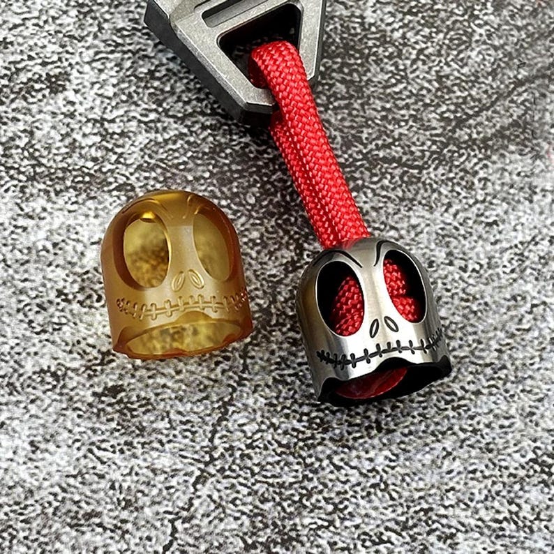 May include: Two keychains featuring a skull design. One keychain is made of clear plastic and the other is made of metal. Both keychains have a red string attached.