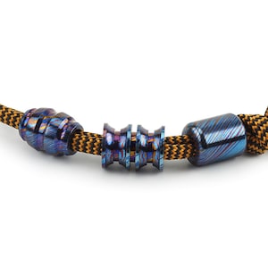 May include: A close-up of a blue and purple patterned metal bead bracelet with a black and yellow cord. The bracelet has three smaller beads and two larger beads.