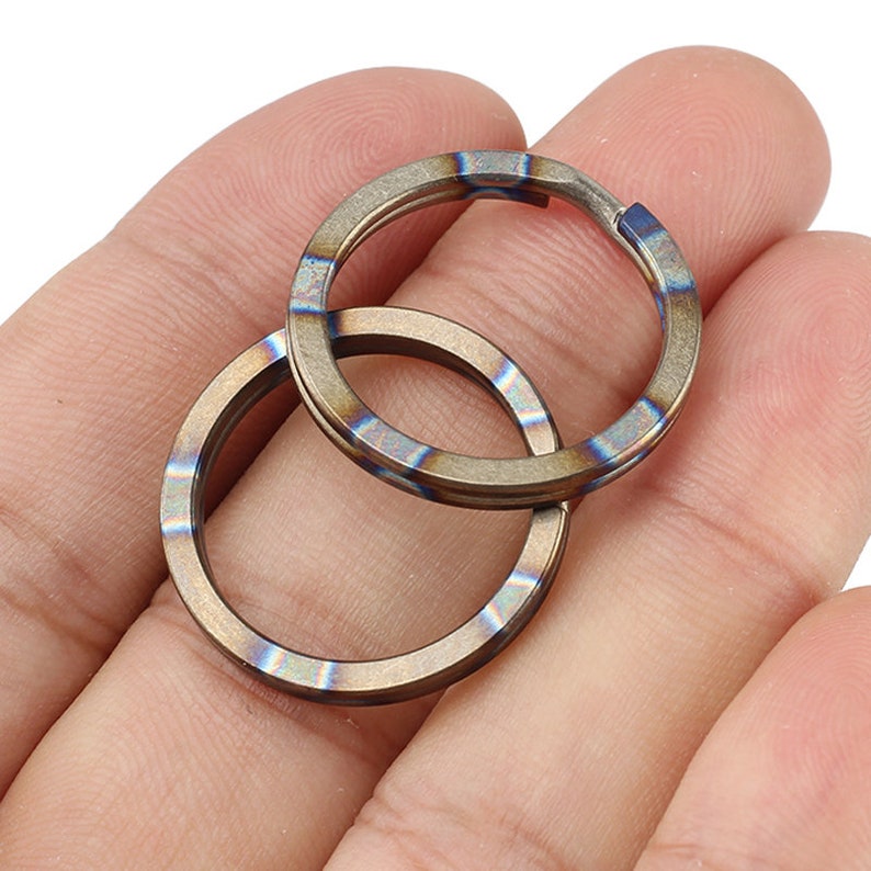 Op de afbeelding: Twee titanium sleutelringen met een blauwe en gouden iriserende afwerking. De ringen zijn in elkaar gehaakt en worden in een hand gehouden.