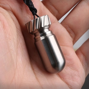 May include: A small, cylindrical, metallic pendant with a brushed finish and a rounded bottom. The top features a gear-like, grooved cap design. A thin black cord is attached to the top of the pendant, allowing it to be worn as a necklace.