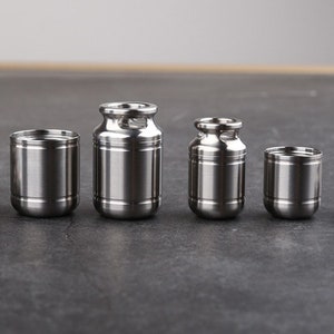 Peut inclure: Quatre récipients en acier inoxydable de tailles différentes sont présentés. Les récipients ont une finition en métal brossé. Deux ont la forme d'un pot à lait avec une poignée. Les deux autres sont cylindriques.