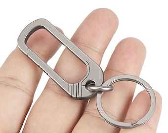 May include: A silver titanium keychain with a large oval-shaped carabiner and a smaller round ring. The carabiner has a textured design and a spring-loaded gate.