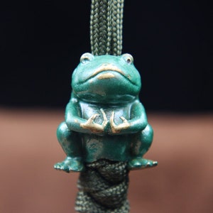 May include: A green frog figurine with gold accents sits in a meditative pose on a green cord. The frog has a serene expression and its hands are folded in its lap.