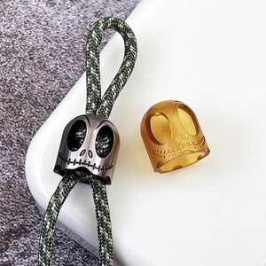 May include: Two skull-shaped charms, one silver and one amber, with a green and white paracord lanyard. The silver charm has a stitched mouth and the amber charm is translucent.