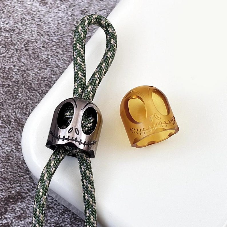 May include: Two skull-shaped charms, one silver and one amber, with a green and white paracord lanyard. The silver charm has a stitched mouth and the amber charm is translucent.