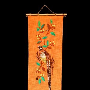 May include: Orange fabric wall hanging with embroidered pheasant and floral design. The pheasant has brown, white, and black details. The embroidery includes green leaves and yellow berries. The top and bottom have bamboo rods.