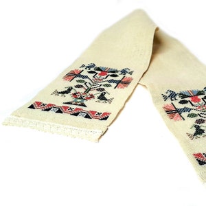 May include: Cream-coloured embroidered scarf with a detailed floral and bird design. The scarf features a repeating pattern of stylized trees, birds, and geometric shapes in red, blue, black, and green. The edges are finished with a delicate lace trim.