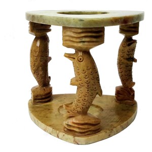 Peut inclure: Un bougeoir décoratif en marbre sculpté à la main. Le support présente trois poissons sculptés soutenant un dessus circulaire. La base est triangulaire. Le marbre est de couleur beige clair avec des veines plus foncées.