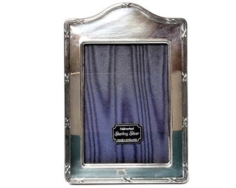 May include: A silver-plated rectangular photo frame with a decorative, curved top edge. The frame has a dark blue backing and a label that reads "Hallmarked Sterling Silver MADE IN ENGLAND."