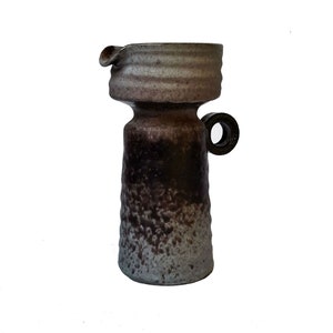 May include: A handmade ceramic jug with a dark brown and grey textured exterior. The jug features a small spout, a handle, and a cylindrical body. The top section is a lighter grey colour.