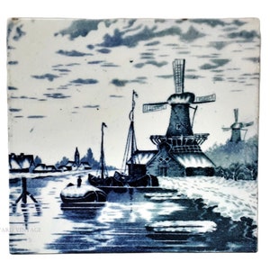 Può includere: Piastrella quadrata in ceramica con una scena paesaggistica blu e bianca. L'immagine raffigura mulini a vento, una barca ed edifici lungo un corso d'acqua. Il cielo è pieno di nuvole. Il design ha un'estetica vintage.