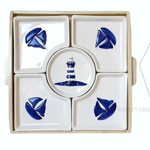 Può includere: Un set da portata a tema nautico con piatti in ceramica bianca decorati con disegni di barche a vela e fari blu. Il set include piatti quadrati e rotondi, disposti in un vassoio di legno con manici. Piatti aggiuntivi sono impilati vicino.
