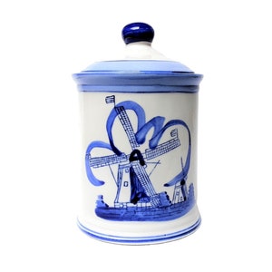 May include: A white ceramic jar with a lid, decorated with a blue and white windmill design. The jar is cylindrical with a lid and a blue knob. The windmill design is painted in blue, with a stylized ribbon-like element.