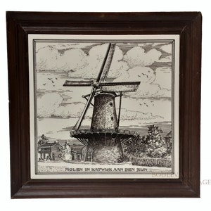 Può includere: Piastrella incorniciata in bianco e nero con una dettagliata illustrazione di un mulino a vento olandese. L'opera d'arte include un cielo con nuvole e uccelli, e il testo "MOLEN IN KATWIJK AAN DEN RIJN". La cornice è marrone scuro.