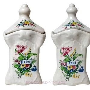 Könnte beinhalten: Zwei weiße Keramik-Gewürzgläser mit Deckel. Jedes Glas hat ein Blumendesign mit rosa, gelben und blauen Blumen und die Wörter "Peper" und "Zout" auf Bannern. Die Gläser haben ein dekoratives, strukturiertes Design.
