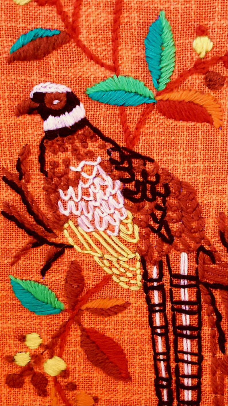 May include: Embroidered bird and foliage design on orange fabric. The bird features brown, white, and yellow details, perched on a branch with green, blue, and orange leaves. The embroidery is detailed and colorful.