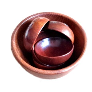 May include: A set of wooden bowls in varying sizes, nested within each other. The bowls are a warm, reddish-brown color, with a smooth, polished finish. The largest bowl is at the bottom, with smaller bowls stacked inside. The wood grain is visible.