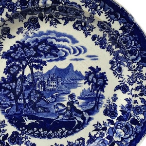Può includere: Piatto in ceramica blu e bianca con una scena paesaggistica dettagliata con un cavaliere a cavallo, un castello e un bordo floreale. Il piatto ha un'estetica classica e vintage.