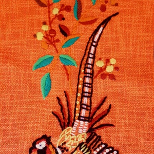 May include: Embroidered pheasant design on orange fabric. The bird features detailed stitching in black, white, red, and yellow. Green and blue leaves and yellow berries accent the design. The background has a textured appearance.