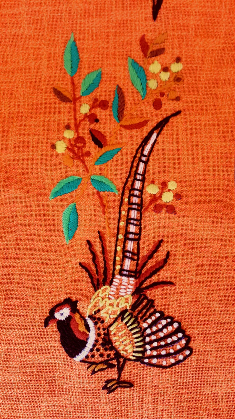 May include: Embroidered pheasant design on orange fabric. The bird features detailed stitching in black, white, red, and yellow. Green and blue leaves and yellow berries accent the design. The background has a textured appearance.