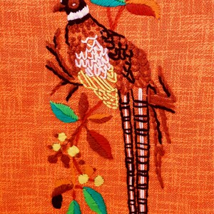 May include: Embroidered pheasant design on orange fabric. The bird is brown, white, and yellow, perched on a branch with green, orange, and yellow leaves. The background is a solid orange color.