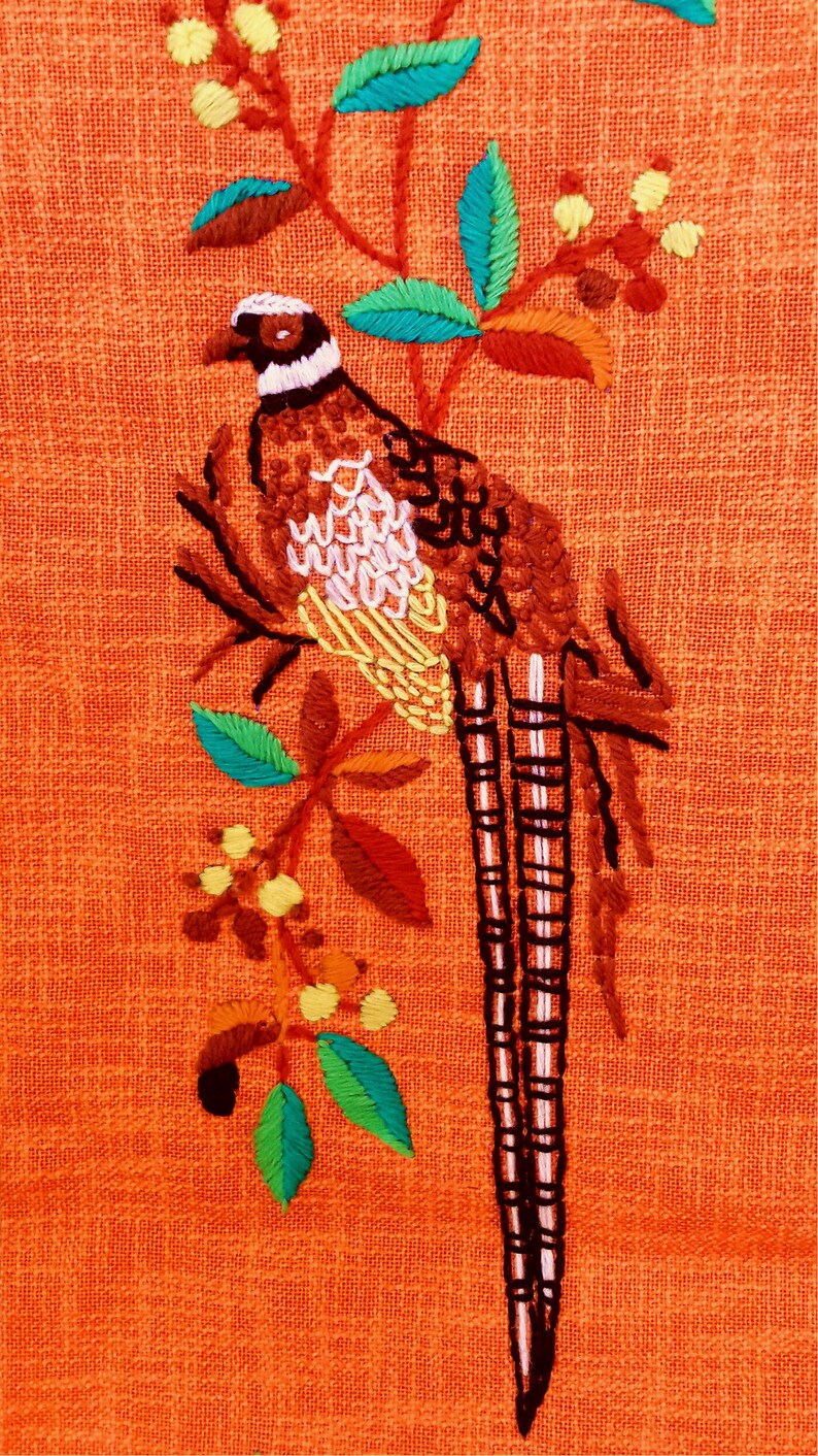 May include: Embroidered pheasant design on orange fabric. The bird is brown, white, and yellow, perched on a branch with green, orange, and yellow leaves. The background is a solid orange color.