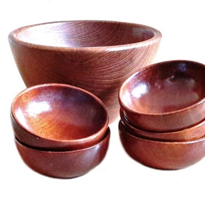 May include: A set of wooden bowls in varying sizes. The largest bowl is deep and round, while the smaller bowls are stacked in groups. The wood has a rich, reddish-brown color and a visible grain pattern, suggesting a natural finish.