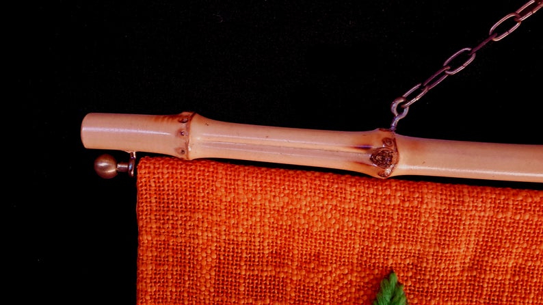 May include: Close-up of a bamboo rod with a metal chain and a hanging orange fabric. The bamboo rod has a natural, light brown color and is attached to the fabric with a metal hook. The fabric has a textured appearance.
