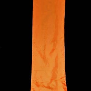 May include: A long, vertical orange fabric banner suspended from a bamboo rod. The banner has a silky, flowing texture and is against a black background. The bamboo rod is a light brown color.