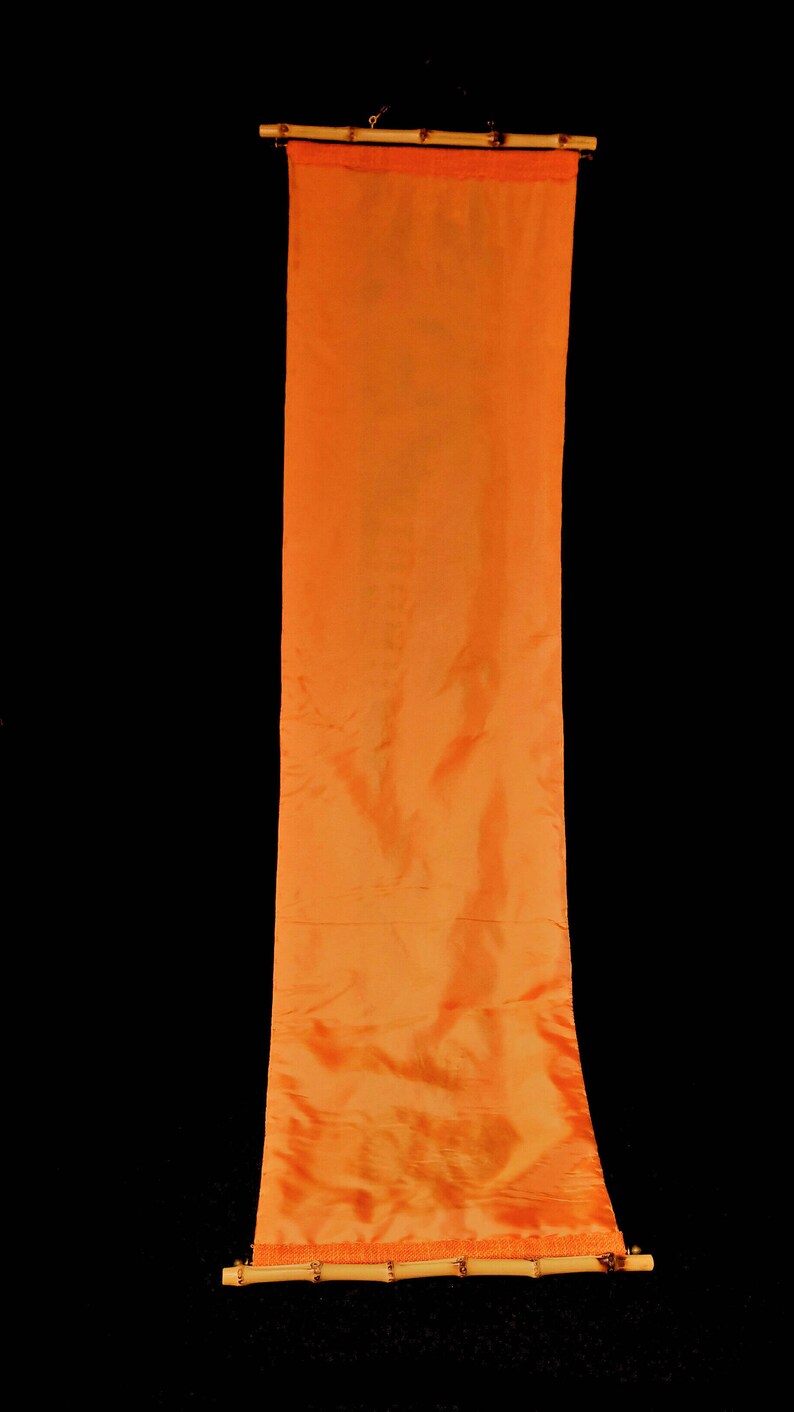 May include: A long, vertical orange fabric banner suspended from a bamboo rod. The banner has a silky, flowing texture and is against a black background. The bamboo rod is a light brown color.
