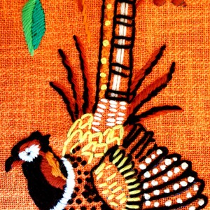 May include: Embroidered pheasant design on orange fabric. The pheasant features black, white, brown, yellow, and orange details. Green leaves and yellow flower accents are also present in the design.