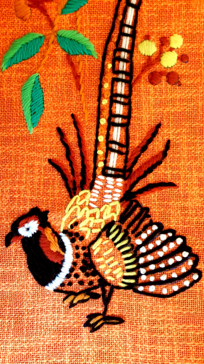 May include: Embroidered pheasant design on orange fabric. The pheasant features black, white, brown, yellow, and orange details. Green leaves and yellow flower accents are also present in the design.